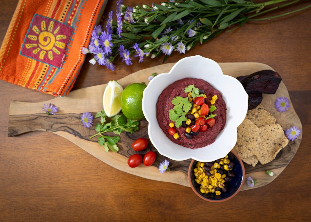 A bowl of black bean beet dip. The bowl is on a decorative wooden board with tomatoes, a lime, a small bowl of corn and beans, some more chips, and some cilantro. To the left there is a napkin and a bundle of purple flowers.