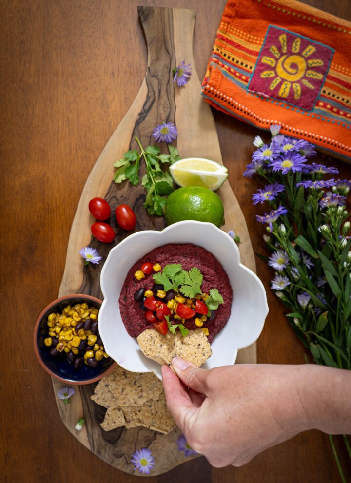 A hand with a chip being dipped into a bowl of black bean beet dip. The bowl is on a decorative wooden board with tomatoes, a lime, a small bowl of corn and beans, some more chips, and some cilantro. To the right there is a napkin with a sun decoration on it and a bundle of purple flowers.