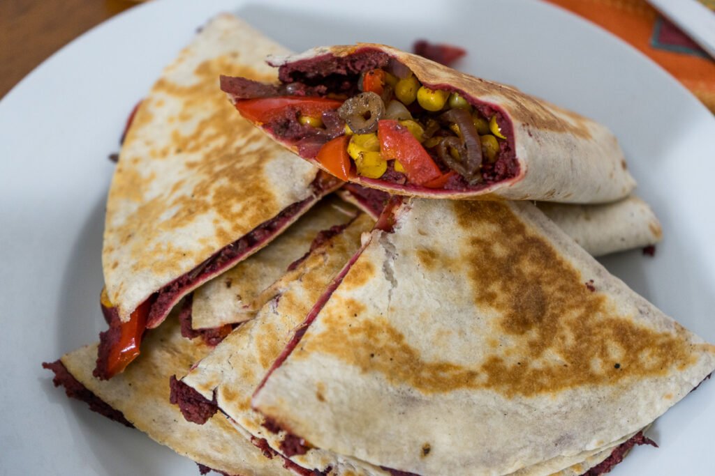 A close up of a pile of cheese-free quesadillas cut into quarter circles on a circular white plate. There are sauteed onions, peppers, and corn along with a bright purple-reddish dip stuffed inside the quesadillas.