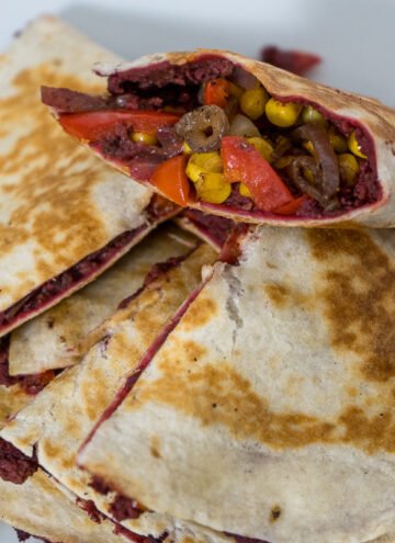 A close up of a pile of cheese-free quesadillas cut into quarter circles on a circular white plate. There are sauteed onions, peppers, and corn along with a bright purple-reddish dip stuffed inside the quesadillas.