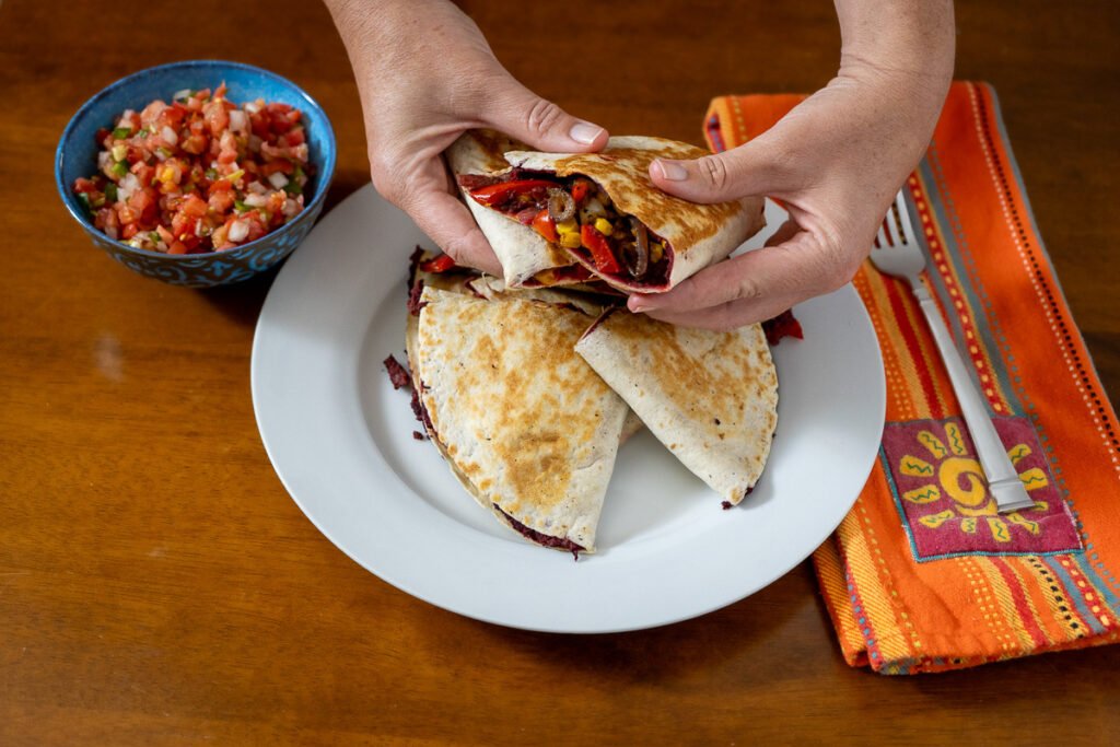 There is a pile of cheese-free quesadillas cut into quarter circles on a circular white plate. A pair of hands is holding two of the pieces of the quesadillas to showcase the delicious looking sauteed veggies inside. There is a blue bowl with salsa in it to the left and a colorful napkin with a fork to the right.