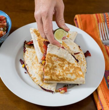 There is a pile of cheese-free quesadillas cut into quarter circles on a circular white plate. A hand is squeezing a lime over the quesadillas. There is a blue bowl with salsa in it to the left and a colorful napkin with a fork to the right.