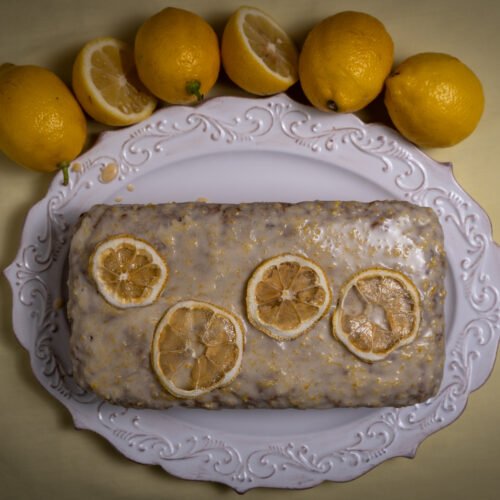 A straight down image of a lemon zucchini cake with white icing and four dehydrated lemon slices on top as decoration. Five lemons surround the plate on the top half of the image and one of the lemons is cut in half. The background is yellow.