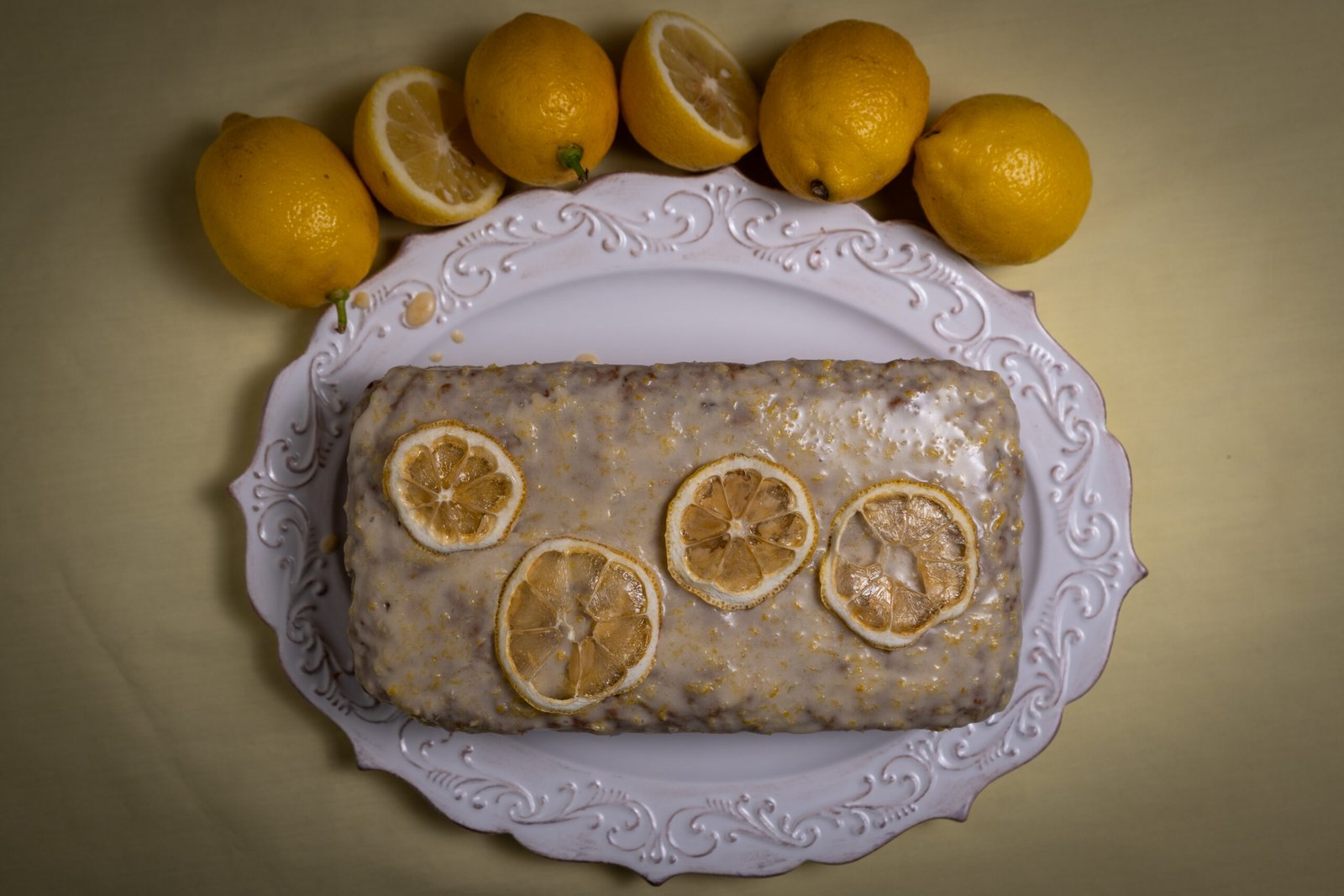 A straight down image of a lemon zucchini cake with white icing and four dehydrated lemon slices on top as decoration. Five lemons surround the plate on the top half of the image and one of the lemons is cut in half. The background is yellow.