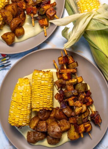 A plate of corn on the cob and seasoned potatoes. There are also several skewers with BBQ tofu, zucchini, and onions. They are laying on a bed of jalapeno creme sauce.