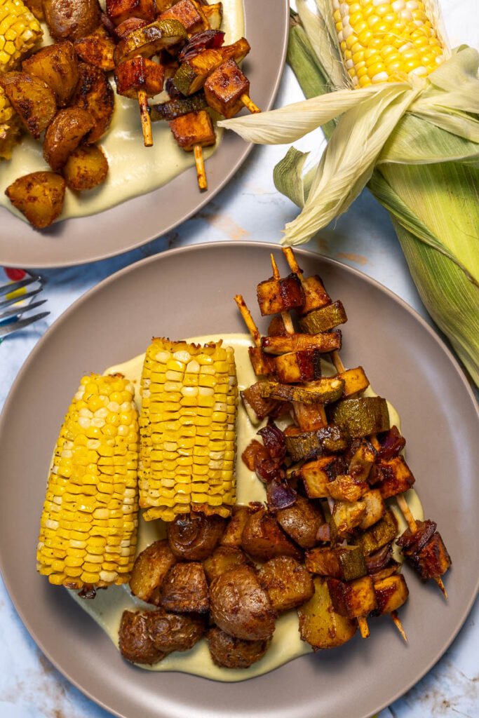 A plate of corn on the cob and seasoned potatoes. There are also several skewers with BBQ tofu, zucchini, and onions. They are laying on a bed of jalapeno creme sauce.