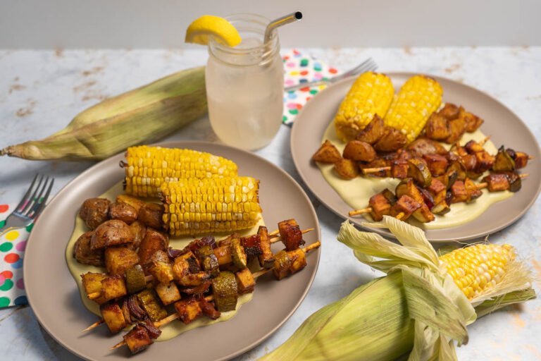 Two plates with corn on the cob, tofu skewers, and potatoes. There are two corn on the cobs with the husks on for decoration, and a Mason Jar with lemonade in it.