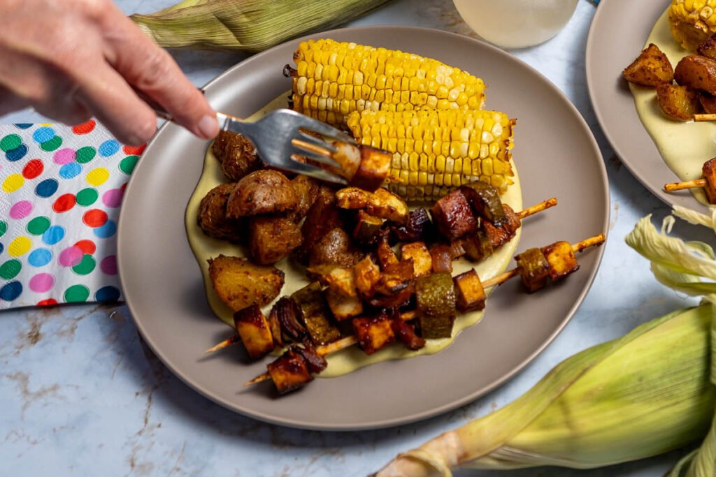 A plate of corn on the cob and seasoned potatoes. There are also several skewers with BBQ tofu, zucchini, and onions. A hand is holding a fork and using the fork to remove the veggies and tofu from the skewer.