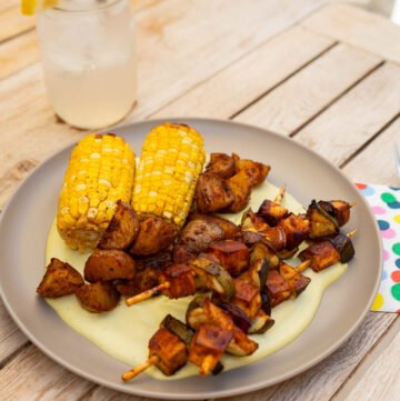 A plate of corn on the cob and seasoned potatoes. There are also several skewers with BBQ tofu, zucchini, and onions. They are laying on a bed of jalapeno creme sauce. There's also a glass of lemonade. The setting is outside on a wooden table.