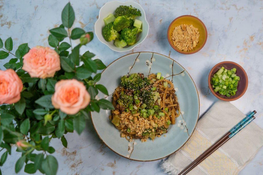 Straight down shot of "beef" and broccoli noodles on a plate. There are pink roses to the left and small bowls with green onions, crushed peanuts and broccoli to the side.