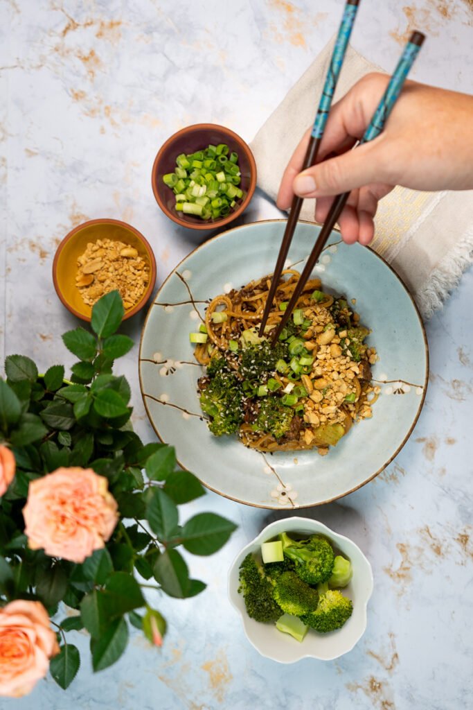 Someone holding chopstick about to eat a plate of five spice vegan "beef" and broccoli noodles .