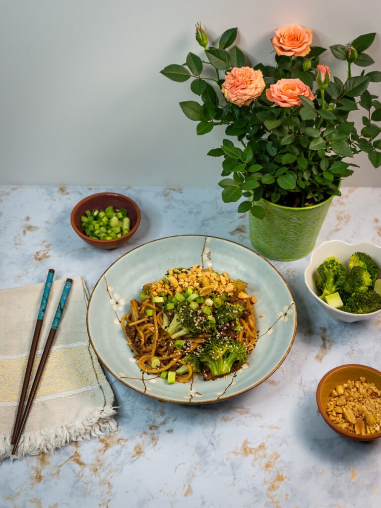 A shot of "beef" and broccoli noodles on a plate. There are pink roses to the right and small bowls with green onions, crushed peanuts and broccoli to the side. There are chopsticks on a white cloth napkin to the right.