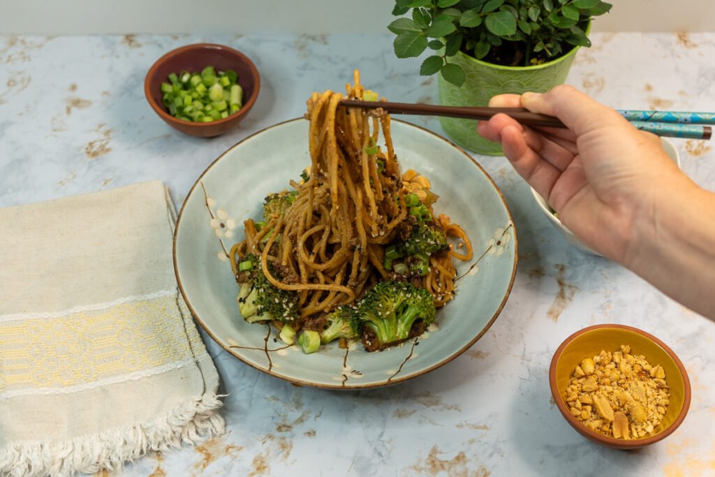 Someone picking up "beef" and broccoli noodles with hand holding chopsticks.