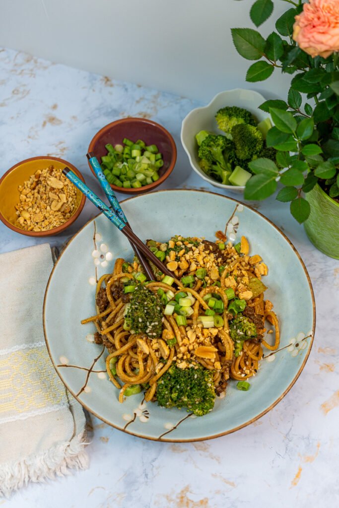 A round blue plate with "beef" and broccoli noodles and chopsticks.