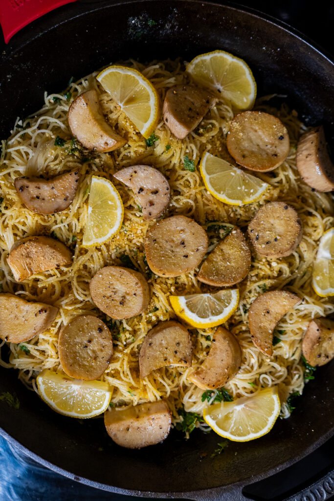 A dish of angel hair pasta, scallops (made out of mushrooms), and lemons in a cast iron pan.