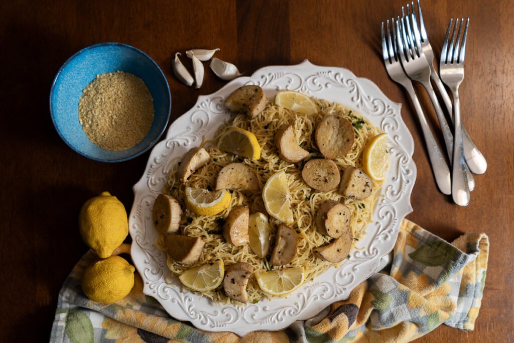 A white plater of angel hair pasta with vegan scallops and lemons. There are a few forks off to the side, a side dish of vegan parmesan in a blue bowl, and some lemon and garlic off to the side.