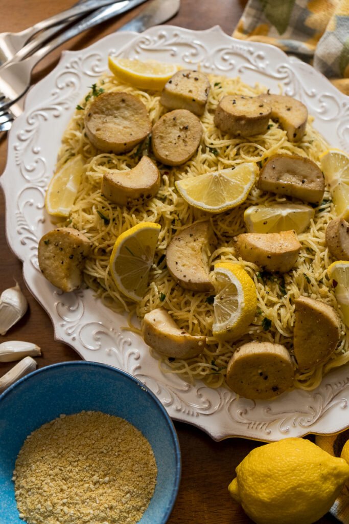 A white plater of angel hair pasta with vegan scallops and lemons. There are a few forks off to the side, a side dish of vegan parmesan in a blue bowl, and some lemon and garlic off to the side.