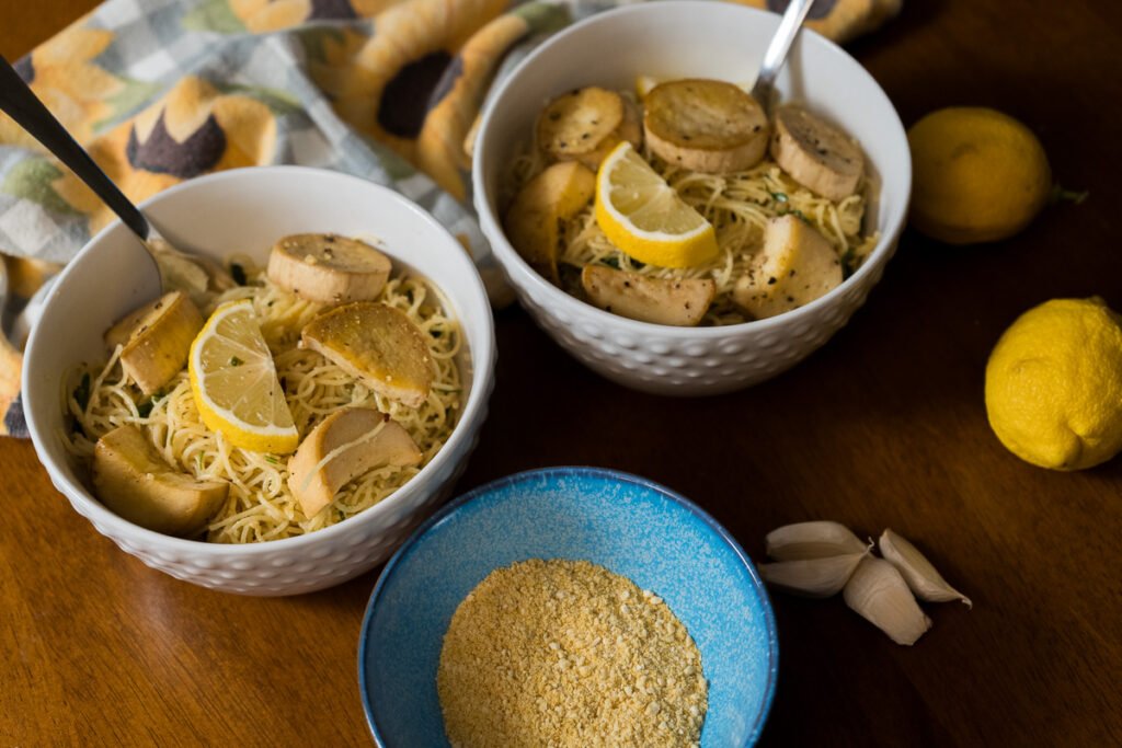 Two white bowls of angel hair pasta and vegan scallops with forks in them. There is also a small blue bowl with vegan parmesan cheese in it.