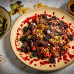 A brown circular plate with Restaurant-style Brussels spouts, vegan feta, and pistachios with a blackberry sauce drizzled on top. On the left there is a bowl of crispy Brussels sprouts leaves and on the right there is a small bowl of blackberries.