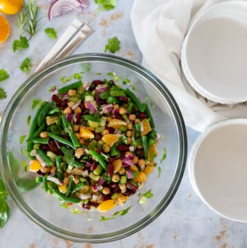 Summer citrus three bean salad with garbanzo beans, kidney beans, green beans, diced onions, herbs, and orange slices in a large clear bowl. There are some smaller serving bowls to the right and some decorative ingredients like oranges and herbs to the left.