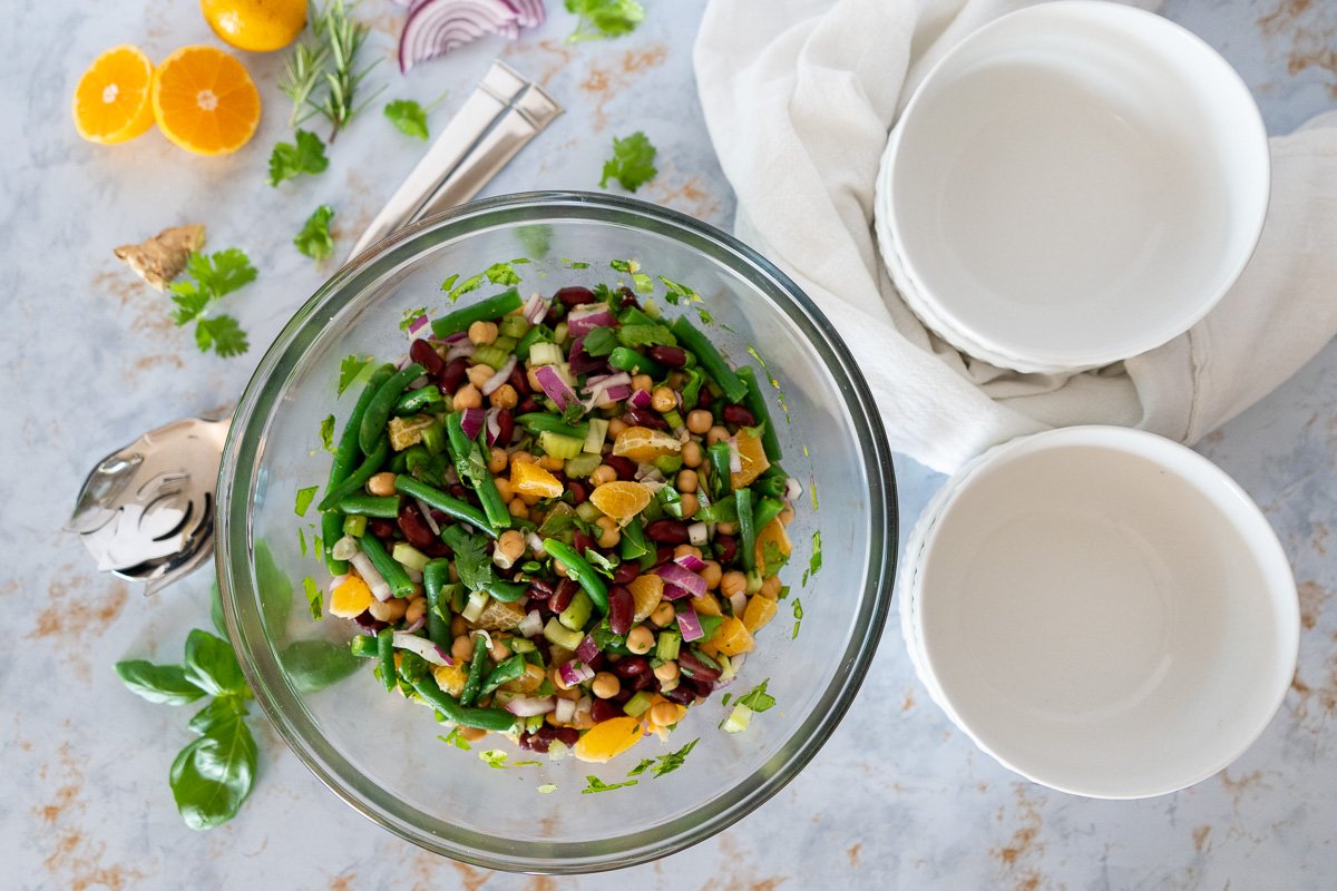 Summer citrus three bean salad with garbanzo beans, kidney beans, green beans, diced onions, herbs, and orange slices in a large clear bowl. There are some smaller serving bowls to the right and some decorative ingredients like oranges and herbs to the left.