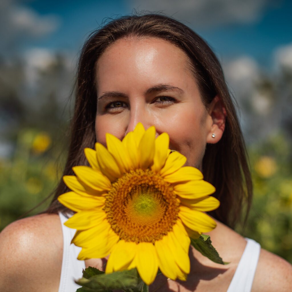 A woman holding a sunflower in front of her face with her mouth covered by the sunflower.