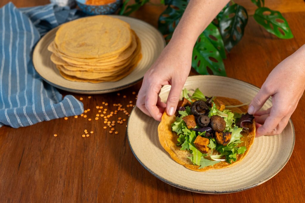 One plate with a large stack of lentil wraps on it to the left, and hand grabbing a lentil wrap with greens and sweet potatoes on a plate to the right.