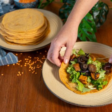 One plate with a large stack of lentil wraps on it to the left, and hand grabbing a lentil wrap with greens and sweet potatoes on a plate to the right.