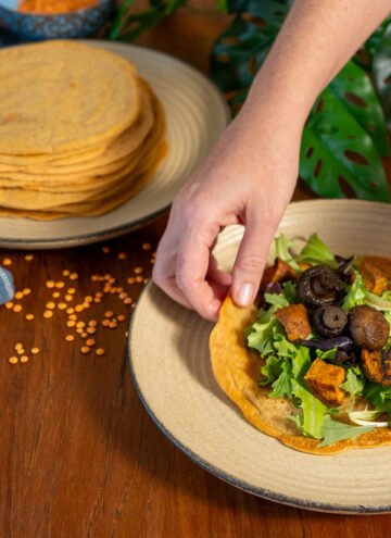 One plate with a large stack of lentil wraps on it to the left, and hand grabbing a lentil wrap with greens and sweet potatoes on a plate to the right.