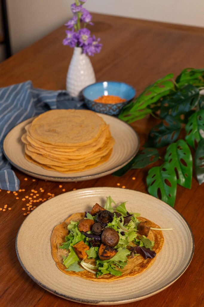 One plate with a large stack of lentil wraps on it and in front of it a plate with a lentil wrap with greens and sweet potatoes on it.