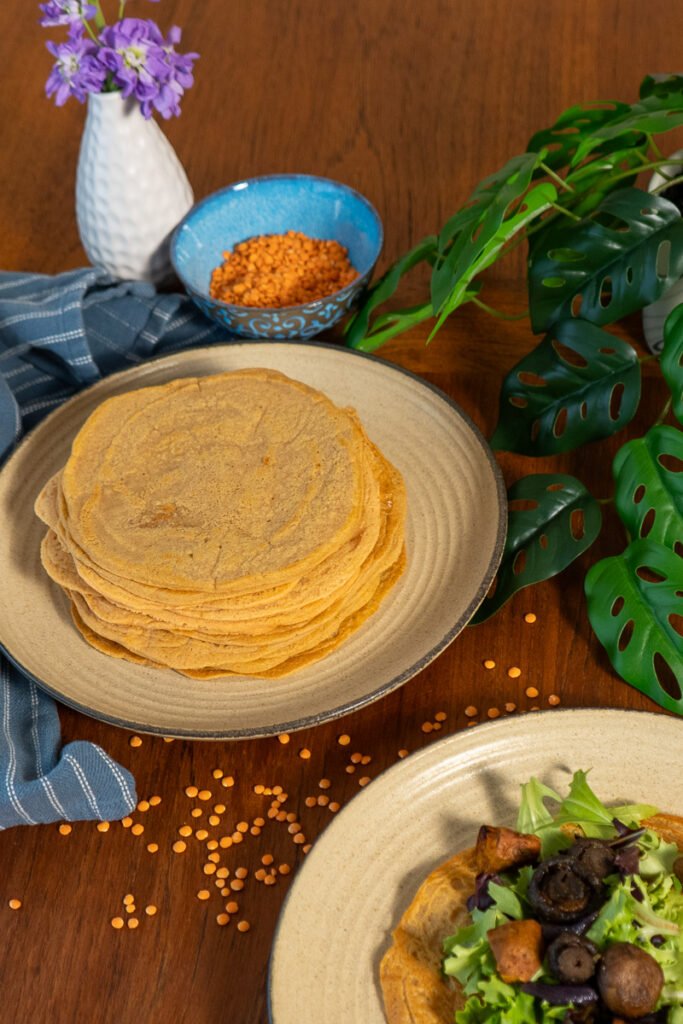 One plate with a large stack of lentil wraps on it and in front of it a plate with a lentil wrap with greens and sweet potatoes on it.