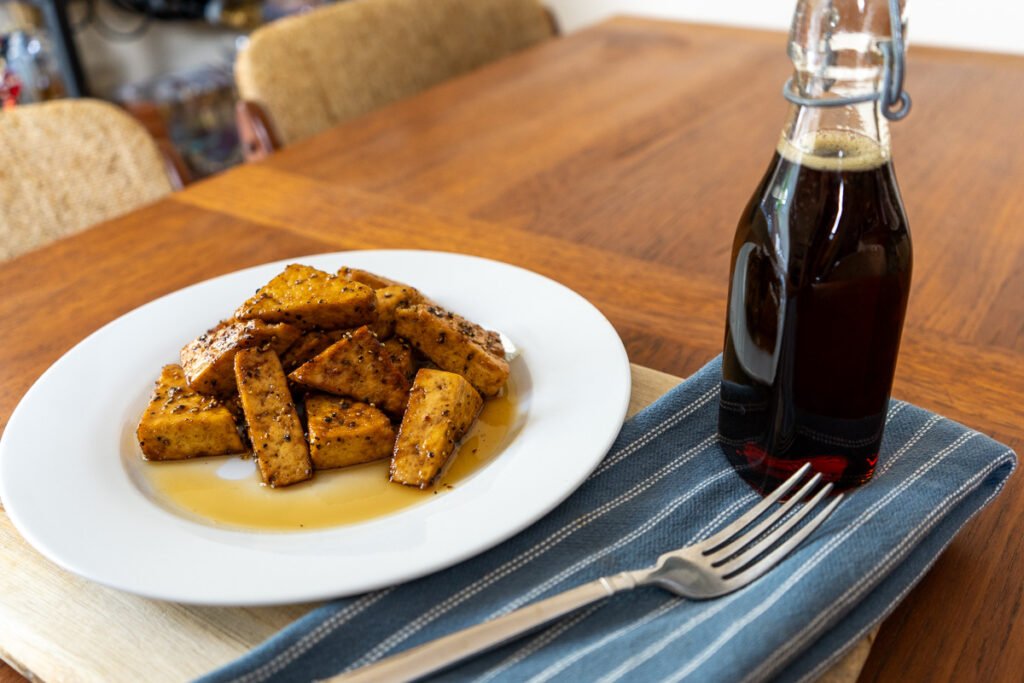 A plate of tofu, a bottle of maple syrup, and a napkin and fork on a table.