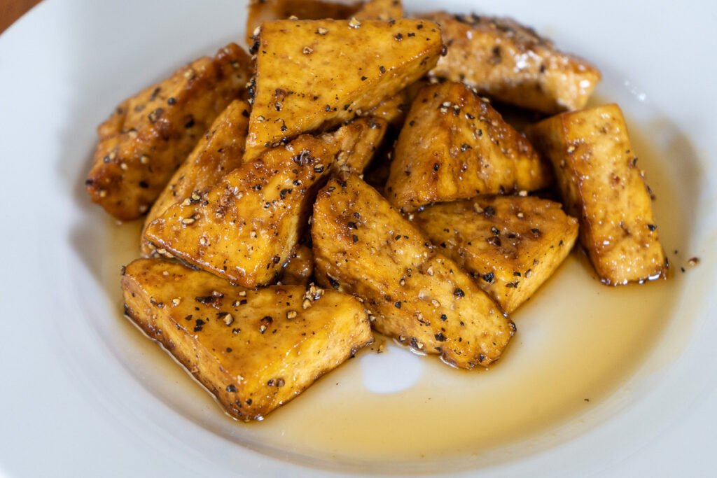 A plate of maple and pepper tofu. The tofu is cut into triangles and stacked in a pile surrounded by maple syrup.