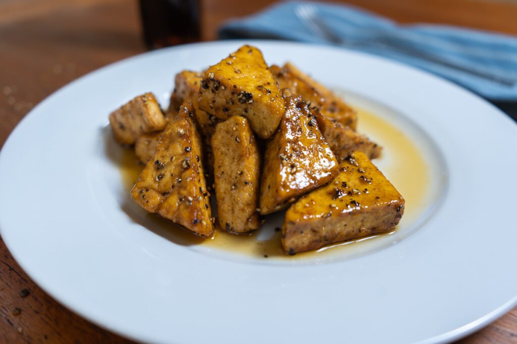 A plate of vegan bacon - maple and pepper tofu. The tofu is cut into triangles and stacked in a pile surrounded by maple syrup.