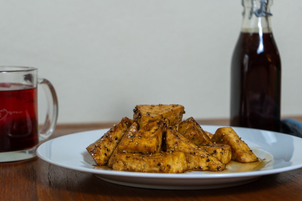 A cup of red tea in a clear glass, a plate of triangle shaped tofu on a white plate and a bottle of maple syrup.