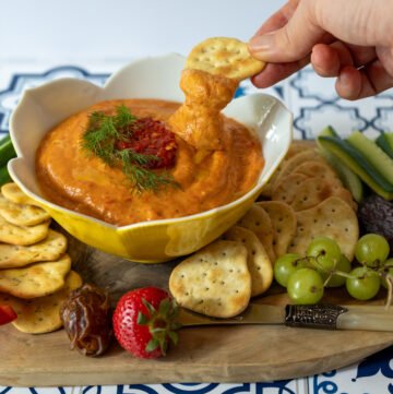 Spicy roasted red pepper dip on a charcuterie board with a hand dipping a cracker into it.