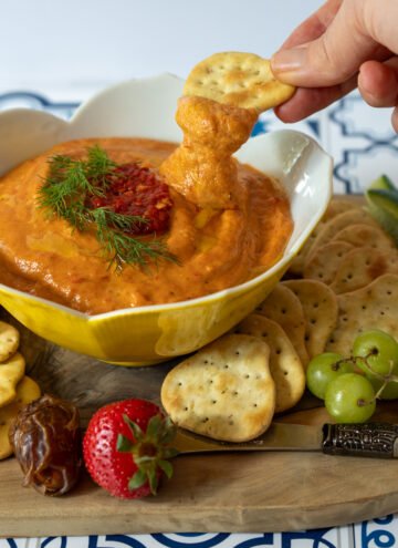 Spicy roasted red pepper dip on a charcuterie board with a hand dipping a cracker into it.