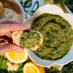 Fingers holding a piece of bread with the kale and herb pesto dolloped on the bread. In the background is a bowl of the kale herb pesto.