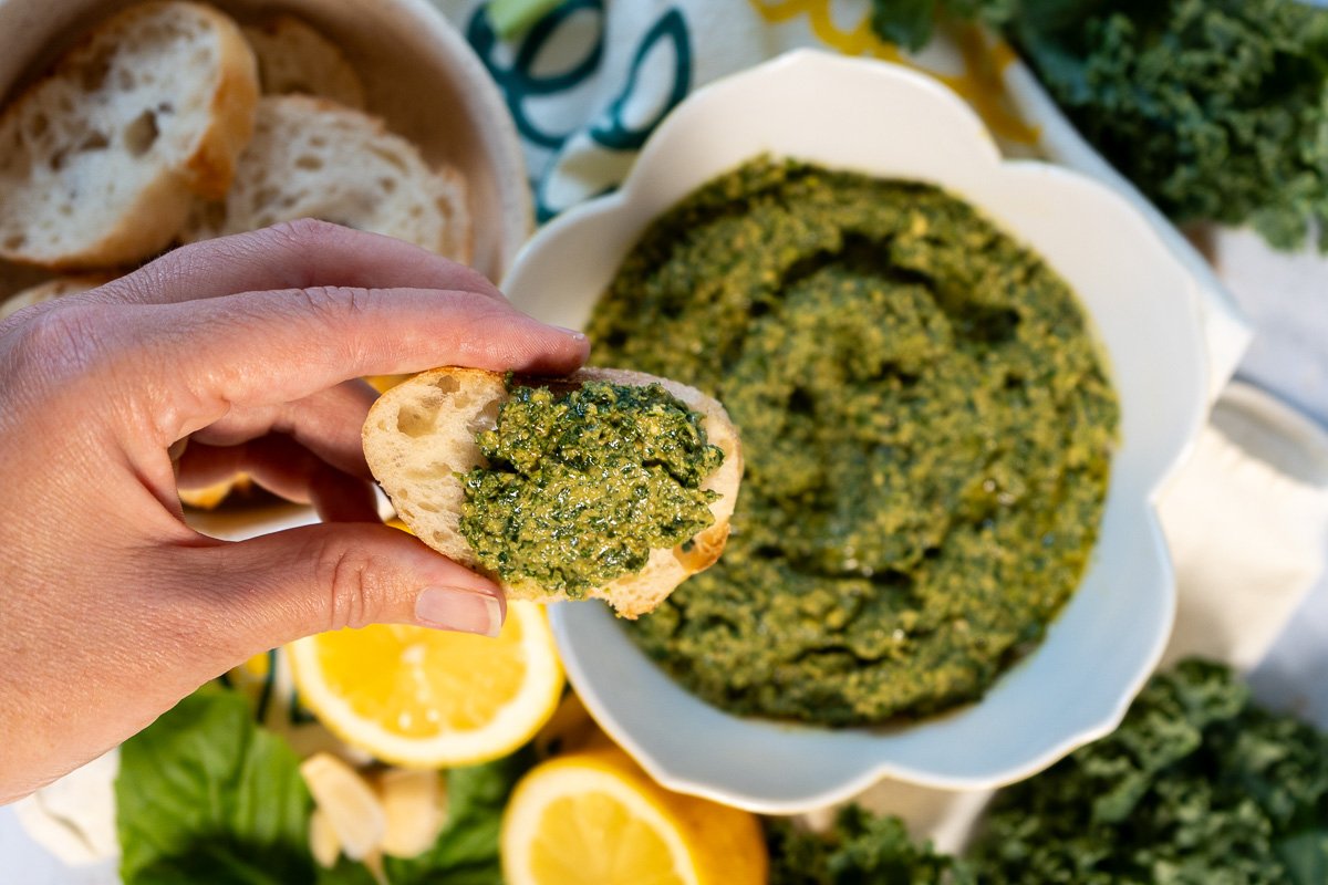 Fingers holding a piece of bread with the kale and herb pesto dolloped on the bread. In the background is a bowl of the kale herb pesto.