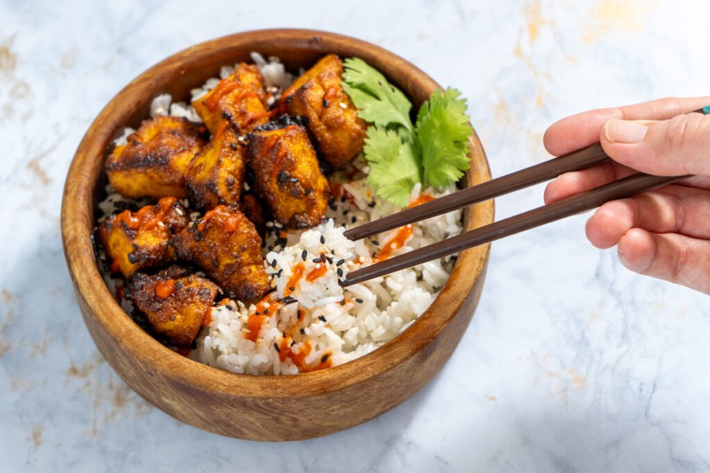 A bowl of coconut rice with Sriracha Tofu on top and hands holding chopsticks to grab some of the rice.