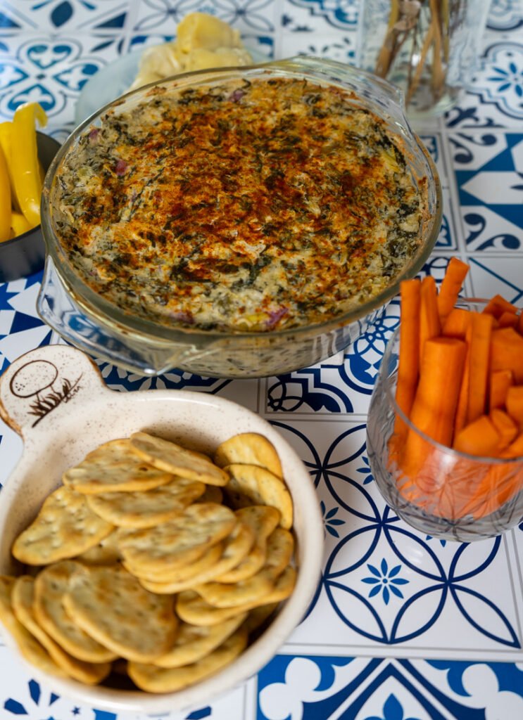 Spinach Artichoke dip in a glass casserole dish with crackers and carrots to the side.