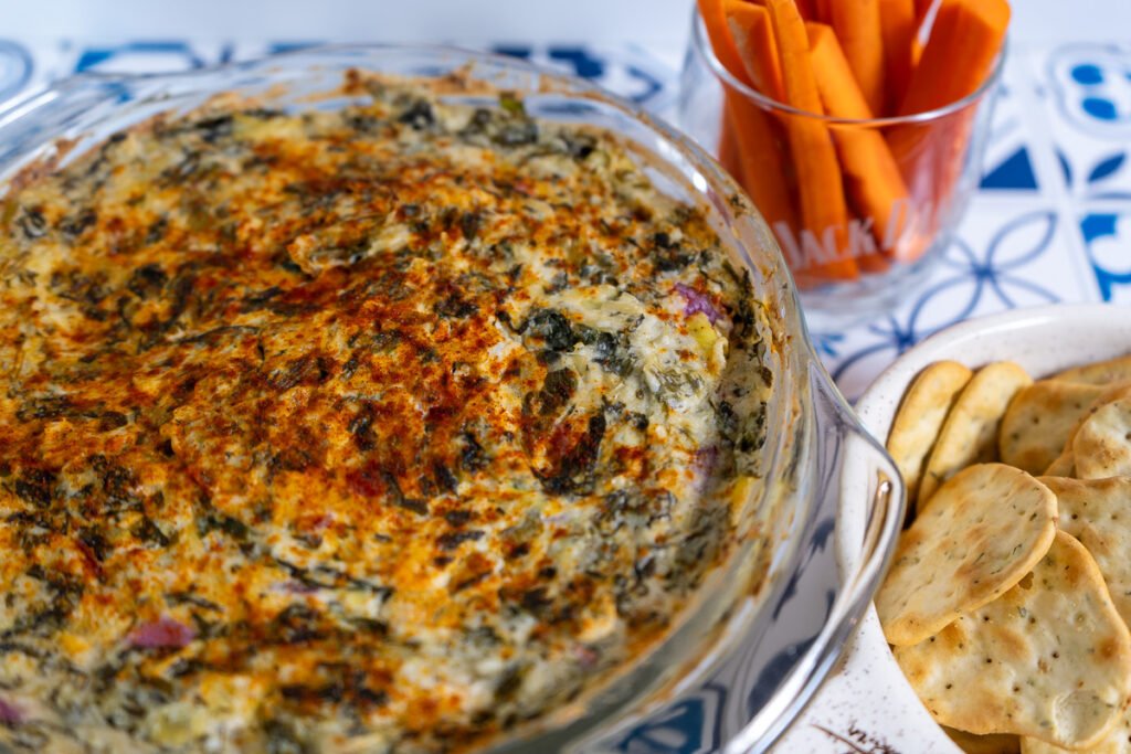 Spinach Artichoke dip in a glass casserole dish with crackers and carrots to the side.