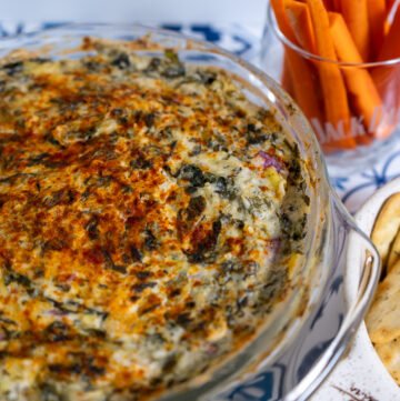 Spinach Artichoke dip in a glass casserole dish with crackers and carrots to the side.