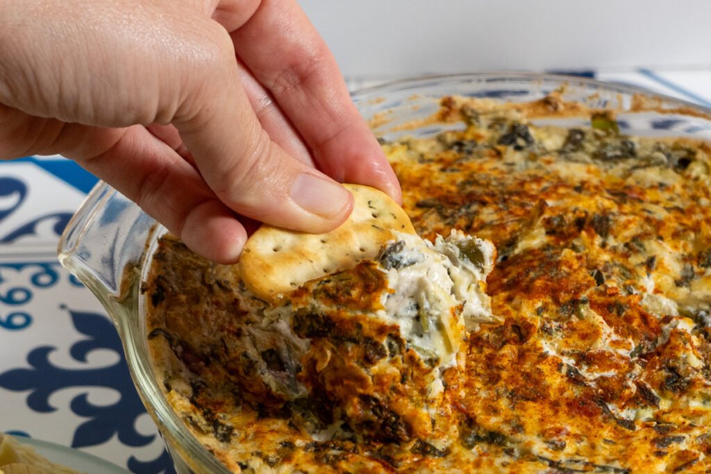 A hand dipping a cracker into the spinach artichoke dip.