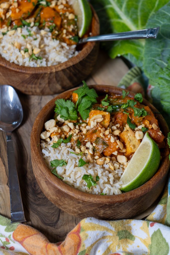 A wooden bowl half filled with brown rice and half filled with West African Tofu Peanut Stew