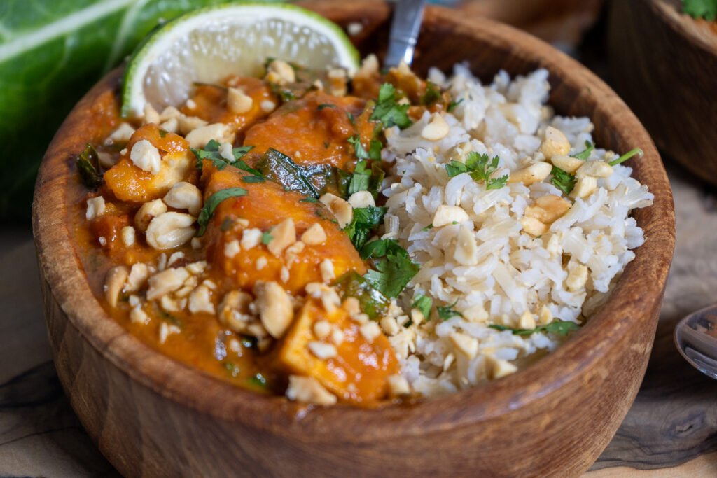 A wooden bowl half filled with brown rice and half filled with West African Tofu Peanut Stew